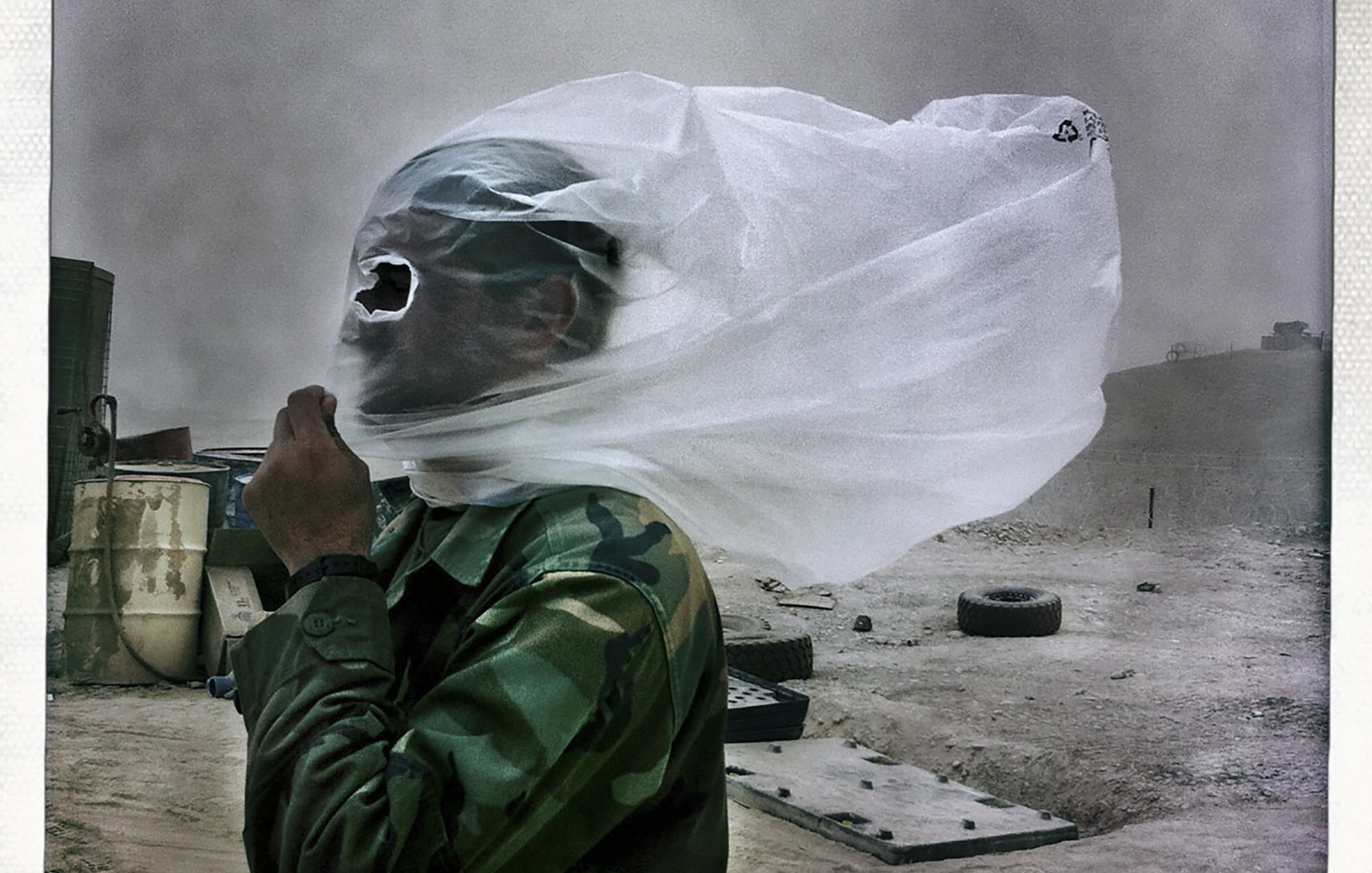 L'image montre un soldat portant un uniforme camouflé, protégé par un morceau de tissu fin qui couvre son visage. Le vent soulève le tissu, créant une atmosphère dramatique. En arrière-plan, le paysage semble poussiéreux et désertique, avec des objets flous tels que des pneus. L'ensemble de la scène évoque un contexte difficile, probablement lié à des conditions climatiques extrêmes.