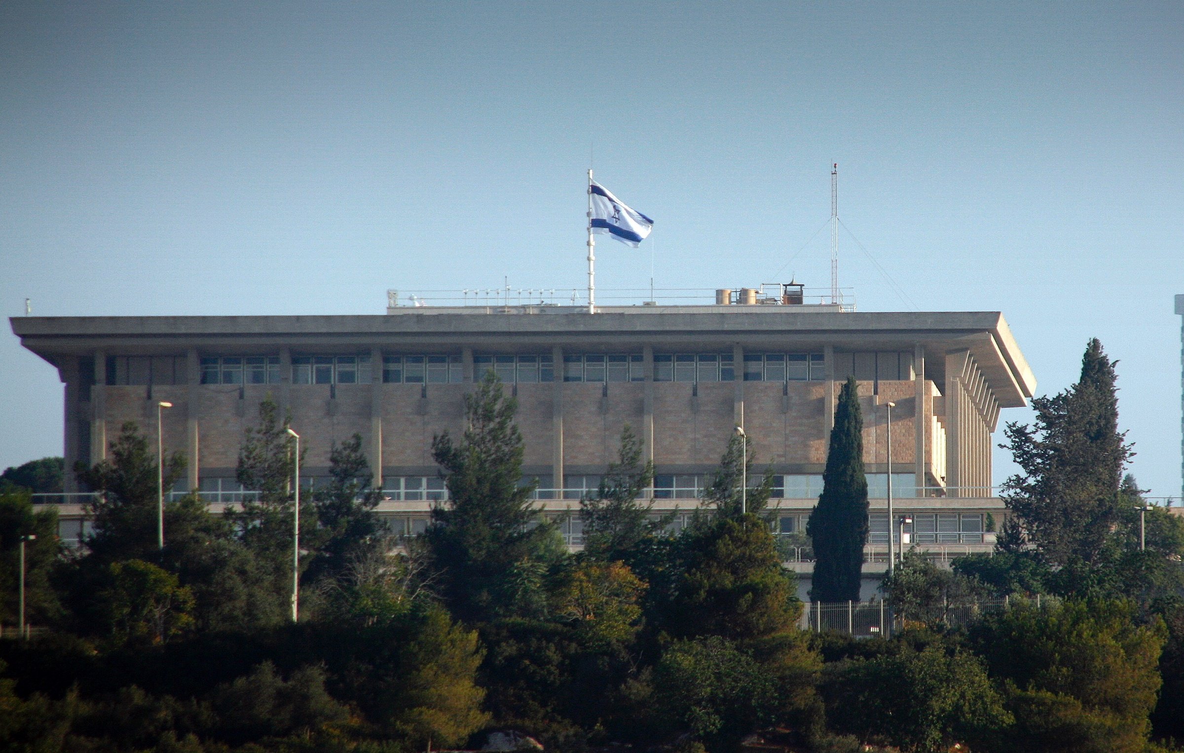 L'image montre un bâtiment imposant, probablement un bâtiment gouvernemental, situé sur une colline. On peut voir le drapeau d'Israël flotter au sommet du bâtiment. Celui-ci est entouré de verdure et d'arbres, indiquant que l'environnement est naturel et paisible. La structure a un style architectural moderne avec de grandes fenêtres et des balcons. L'éclairage suggère que la photo a été prise pendant la journée.