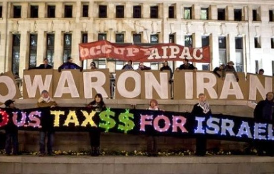L'image montre un groupe de personnes réunies autour d'une grande pancarte ou d'un affichage, exprimant des revendications politiques. Ils tiennent des panneaux où l'on peut lire "NO WAR ON IRAN" et "NO US TAX$$ FOR ISRAEL". L'ambiance semble être celle d'une manifestation ou d'un rassemblement en faveur de la paix et critiquant l'utilisation des fonds publics. L'arrière-plan comprend un bâtiment, suggérant que l'action a lieu dans un espace public.