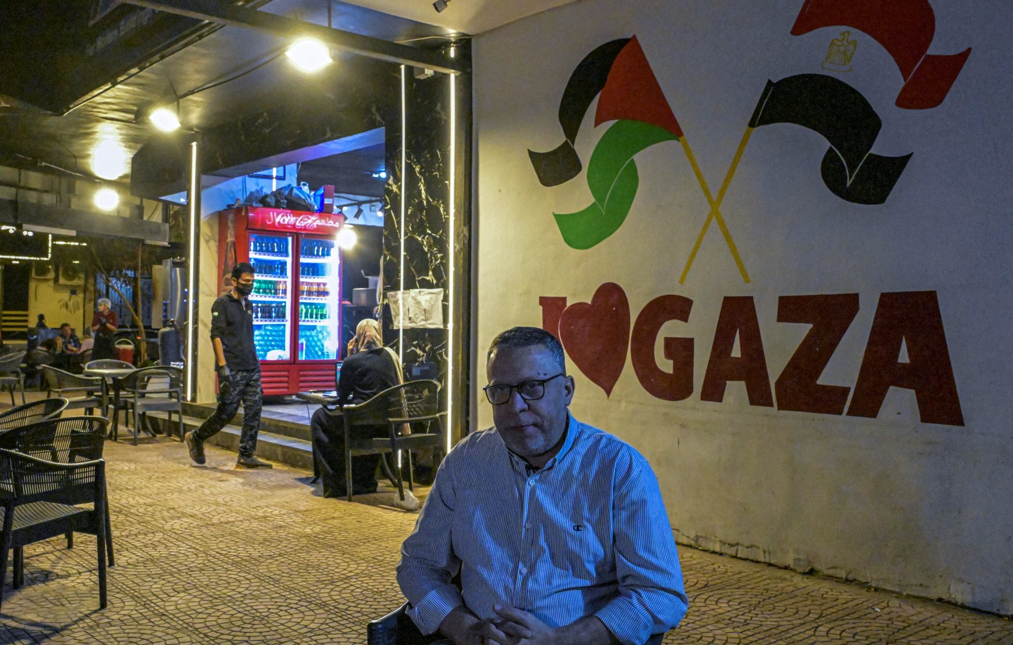 Un homme assis, devant un mural "I ❤️ GAZA", avec des drapeaux palestiniens en arrière-plan.