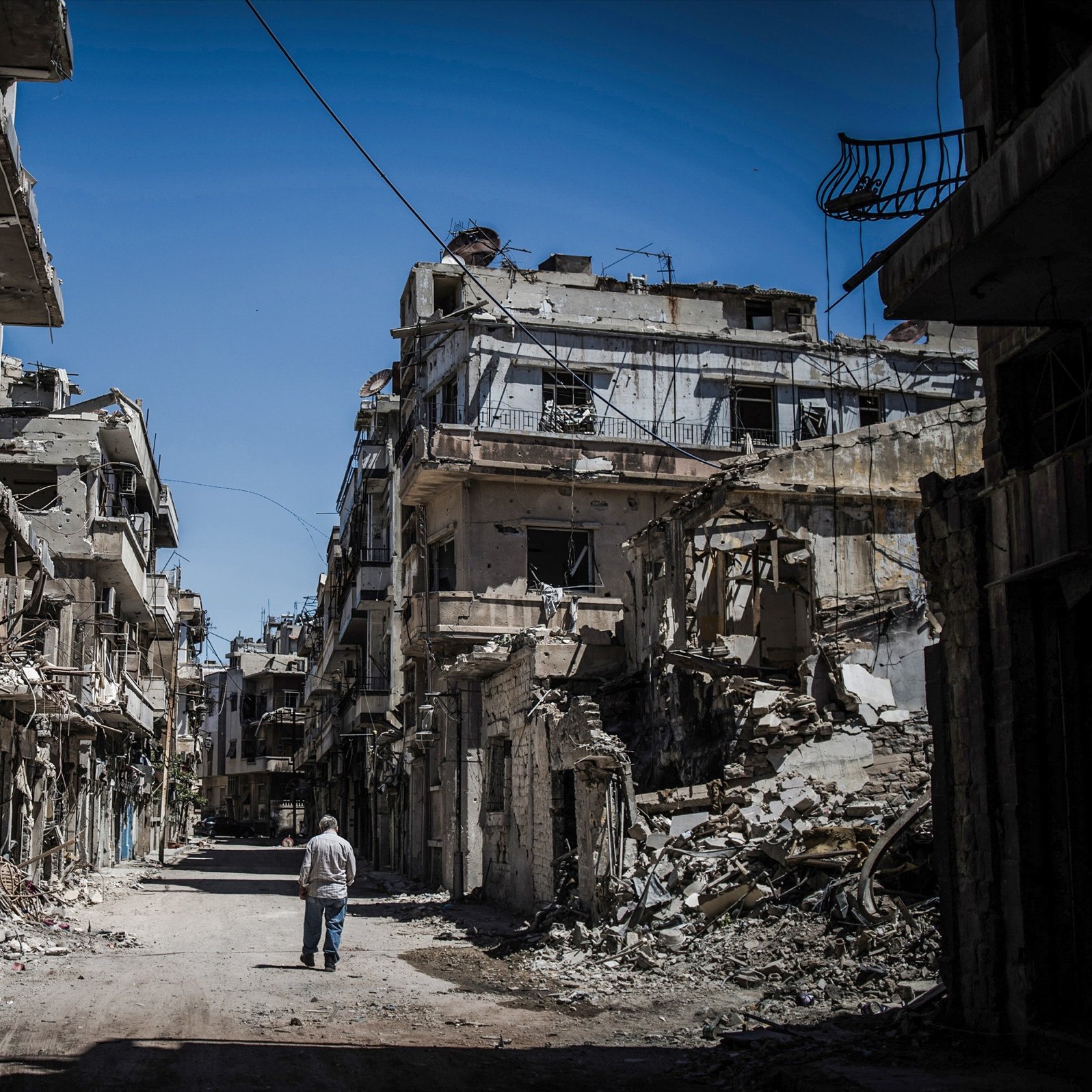 L'image montre une rue dévastée, bordée de bâtiments en ruine. Les murs sont fissurés et effondrés, témoignant des destructions causées par un conflit. Au centre de la scène, un homme marche seul, semblant contempler les vestiges de ce qui était autrefois un quartier vivant. Le ciel est clair, contrastant avec la désolation environnante. Cette image évoque une atmosphère de solitude et de tristesse face à la perte et à la destruction.