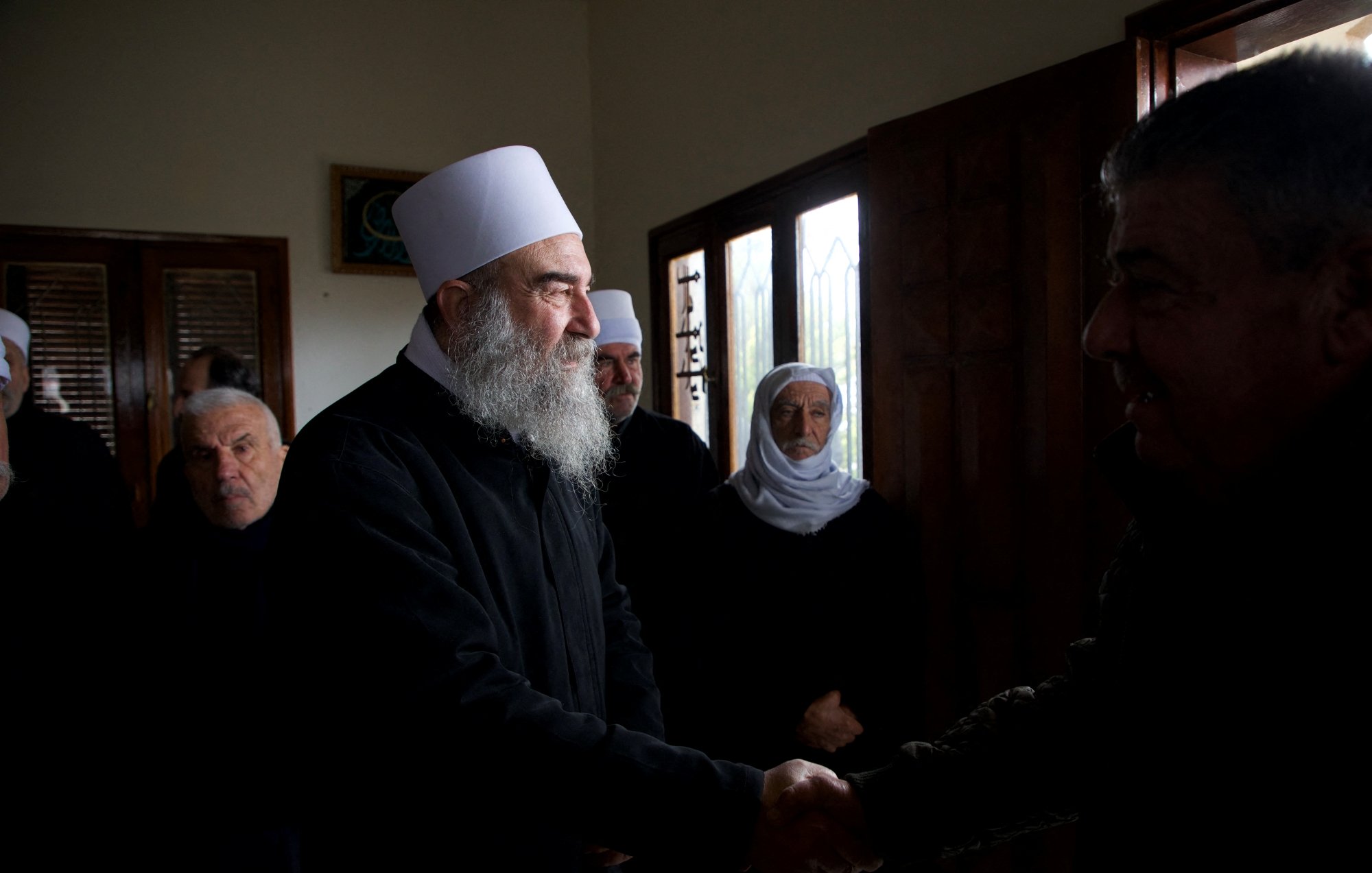 Group of men, some with white hats and beards, shaking hands and wearing dark clothing in a room with windows.