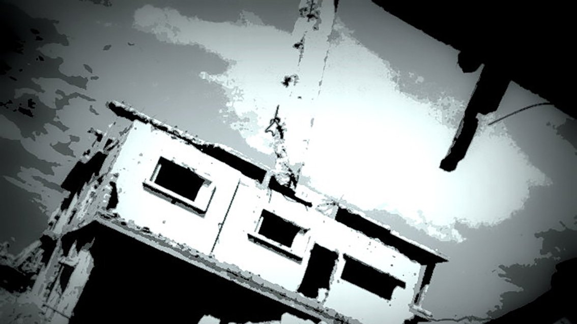 L'image montre un bâtiment en ruine, partiellement détruit, avec des murs endommagés et une atmosphère sombre. La structure semble en déséquilibre, suspendue ou penchée, ce qui renforce l'impression de dévastation. Le ciel est nuageux, ajoutant une tonalité dramatique à la scène. Les teintes sont principalement en noir et blanc, créant un effet visuel frappant.