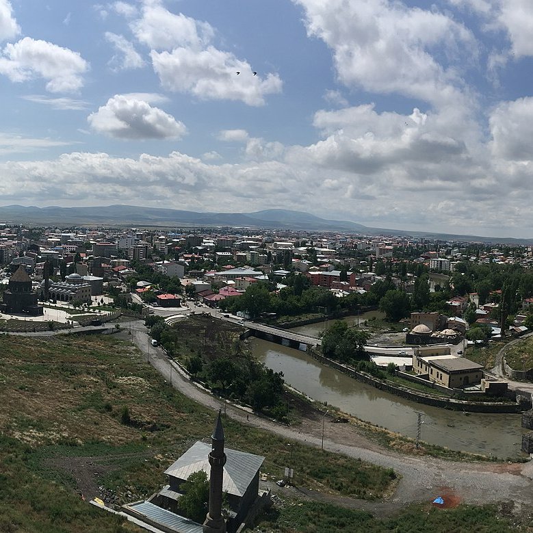 L'image représente une vue panoramique d'une ville avec un paysage verdoyant. On peut voir des toits de maisons, des bâtiments et des infrastructures qui s'étendent à travers la vallée. Le ciel est partiellement nuageux, laissant passer la lumière du soleil. Au premier plan, il y a des collines et des routes qui serpentent à travers le paysage. L'ensemble dégage une atmosphère paisible et naturelle, avec une harmonie entre l'urbanité et la nature environnante.