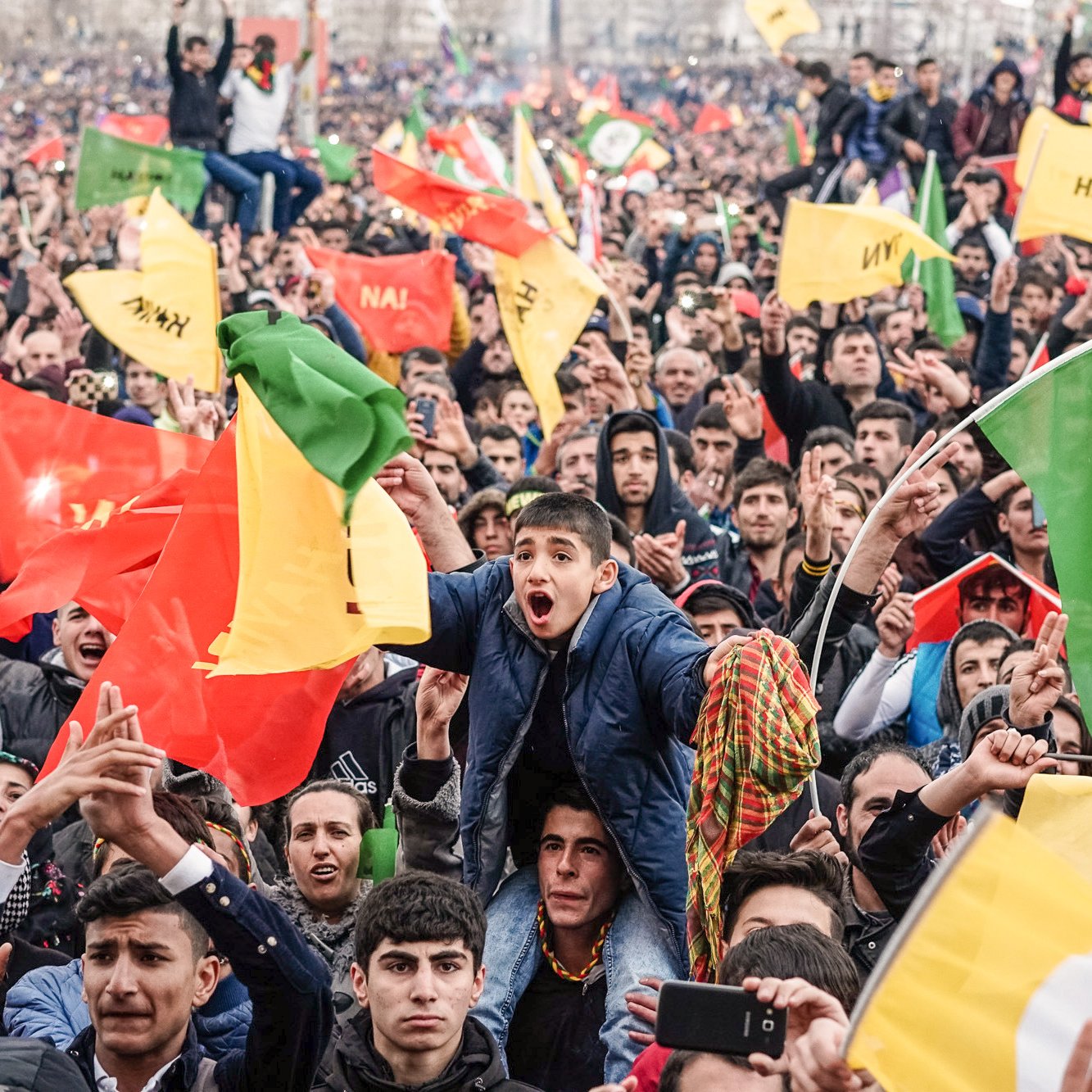 L'image montre une grande foule rassemblée, manifestant avec enthousiasme. Les gens lèvent des drapeaux colorés, dont certains semblent être aux couleurs rouge, jaune et vert. On peut voir un jeune garçon au premier plan exprimant sa joie et son excitation. L'atmosphère est festive, et les participants semblent engagés dans une cause commune, remplis d'énergie et de détermination.