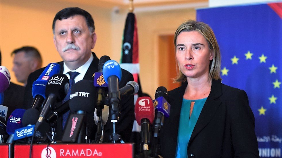 L'image montre deux personnes debout derrière un podium avec plusieurs microphones. La femme, à droite, porte une veste noire et une blouse turquoise, et semble s'exprimer devant les médias. L'homme, à gauche, est également en costume et semble écouter. En arrière-plan, on peut voir des drapeaux et un affichage qui indique une conférence ou un événement officiel. L'ambiance semble être celle d'une conférence de presse.