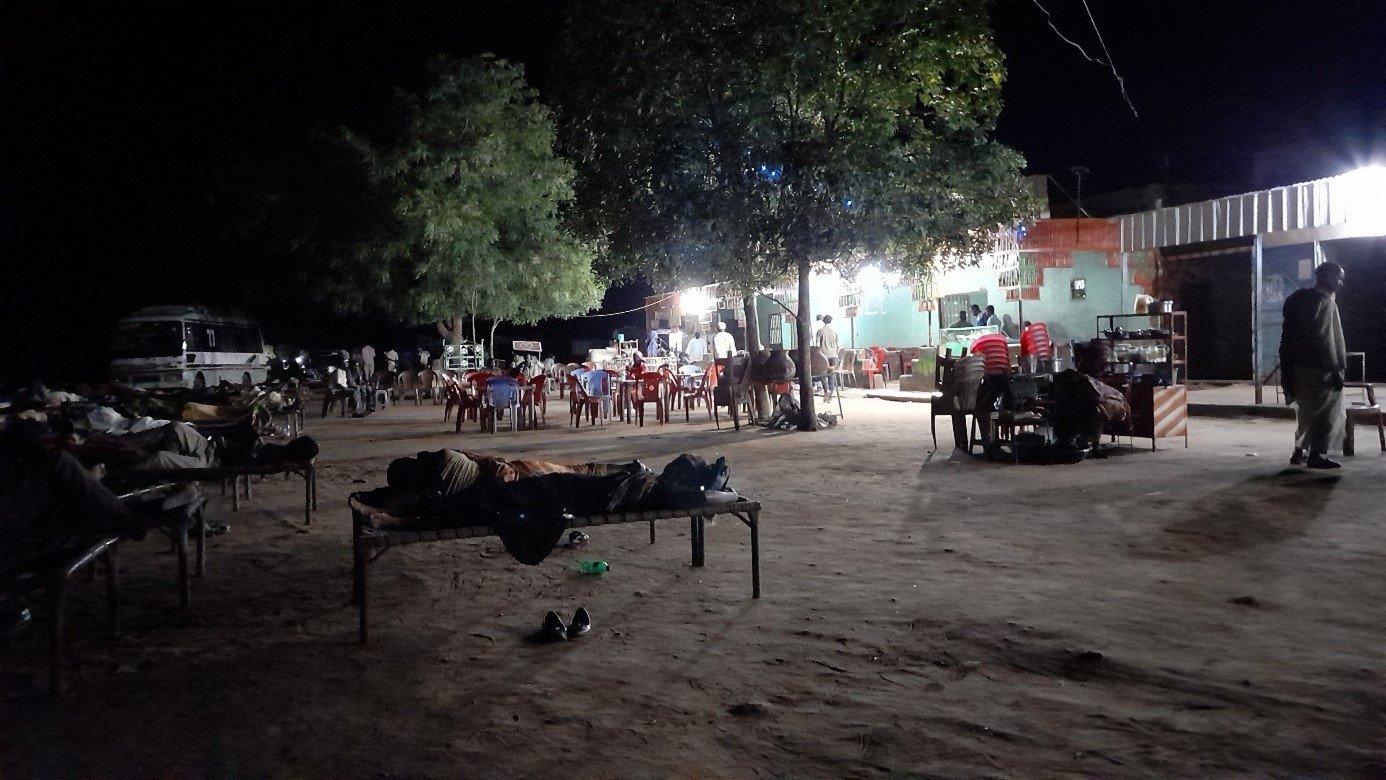 L'image montre un espace en plein air, probablement dans un endroit rural. Il fait nuit, et l'éclairage est principalement fourni par une source lumineuse sur le bâtiment à droite. On peut voir plusieurs tables et chaises disposées, avec des personnes assises et d'autres qui se déplacent. À gauche, il y a des lits ou des matelas disposés sur le sol, suggérant que des gens y dorment ou se reposent. Des arbres sont visibles, ajoutant une touche de verdure à la scène. L'atmosphère semble calme, mais animée par la présence de plusieurs personnes.