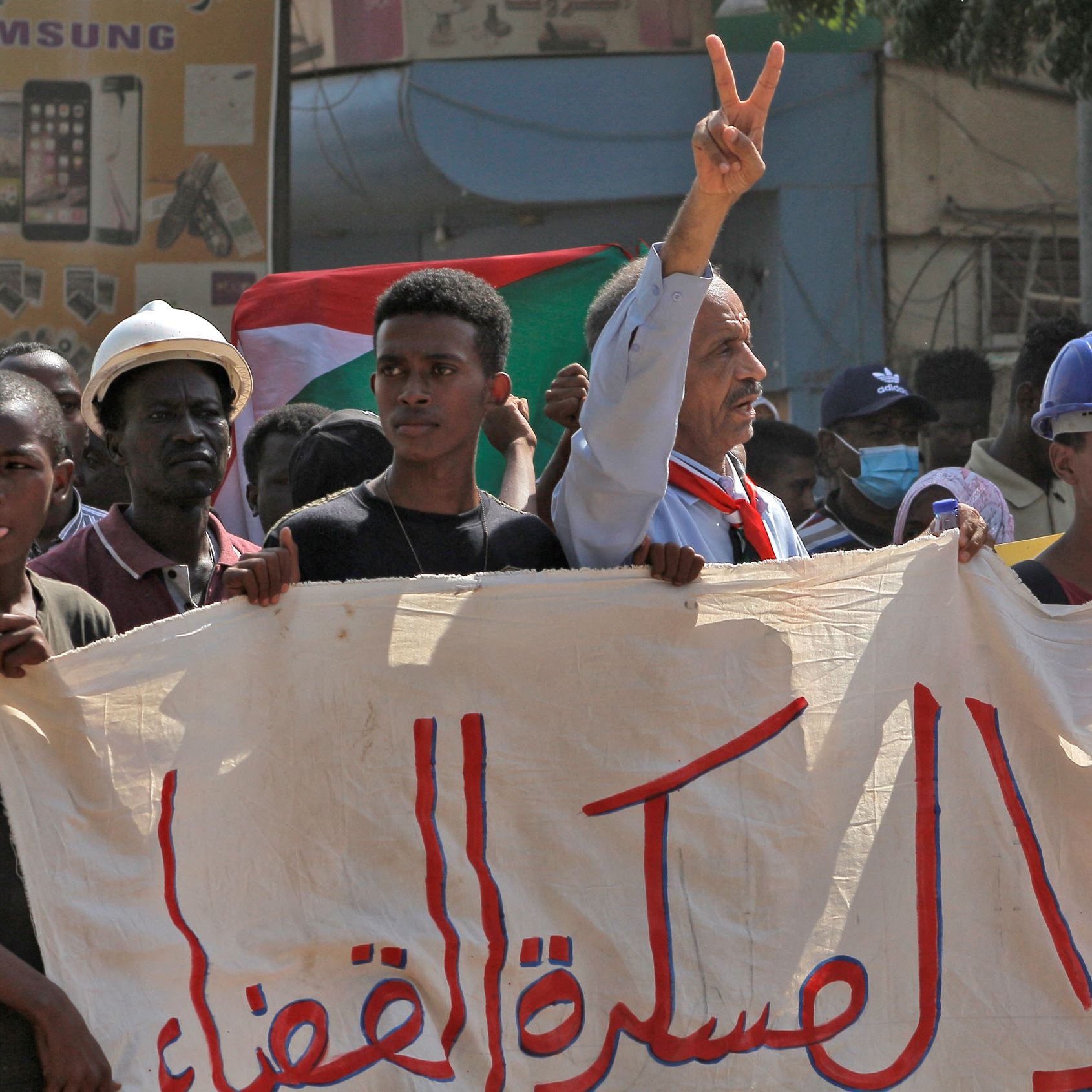 L'image montre une scène de manifestation, probablement en rapport avec des revendications politiques. On y voit des personnes tenant une grande bannière. Un homme au premier plan fait un signe de paix avec ses doigts. Les manifestants semblent déterminés et engagés, et des drapeaux sont visibles en arrière-plan, indiquant un contexte national. L'atmosphère est chargée d'émotion, illustrant un moment de solidarité.