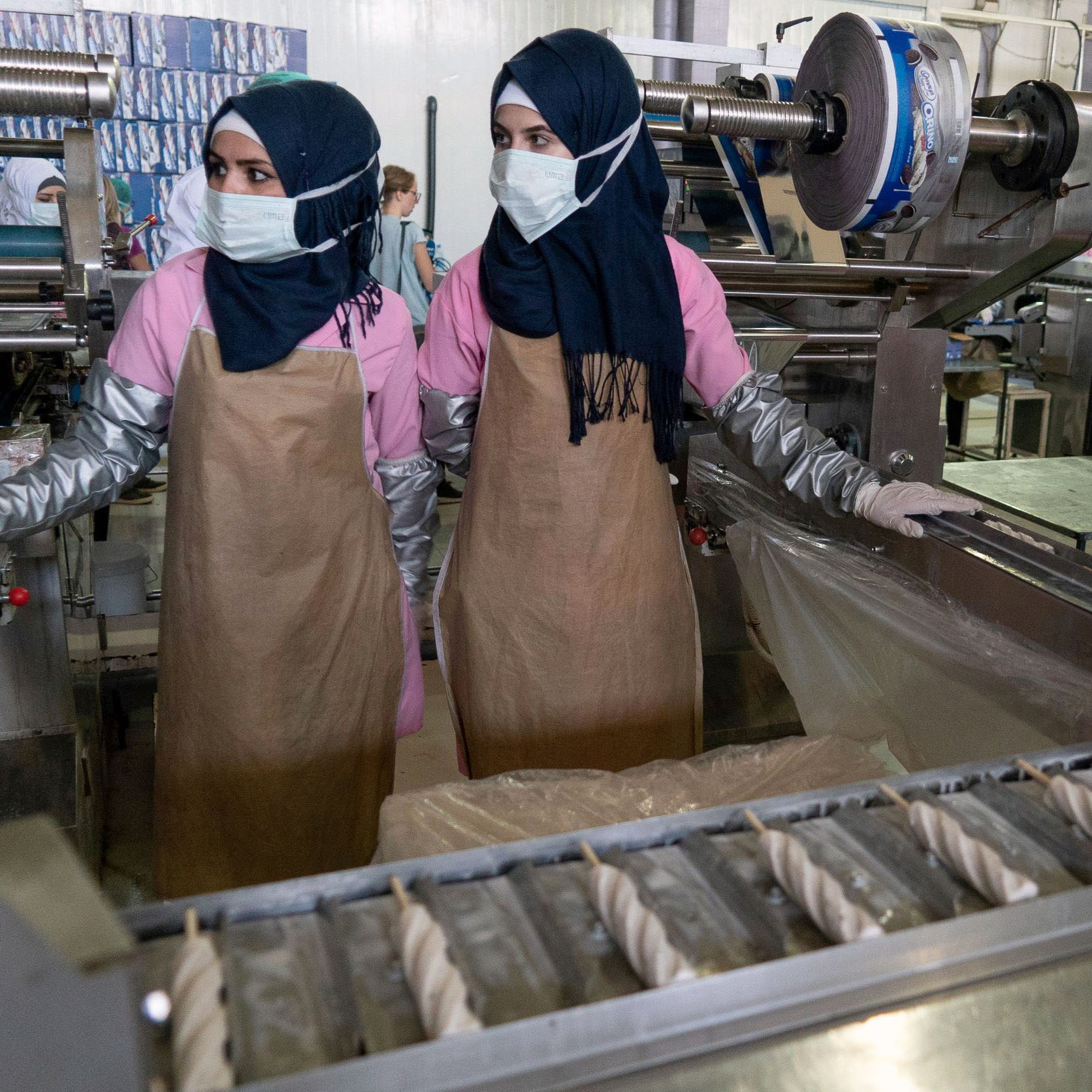 L'image montre deux femmes travaillant dans un environnement industriel. Elles portent des hijabs et des masques de protection. Elles sont habillées de vêtements de travail, avec des blouses et des tabliers. À l'arrière-plan, on peut voir des machines et d'autres personnes également en train de travailler, ce qui indique qu'il s'agit probablement d'une usine ou d'un atelier de production. L'atmosphère semble axée sur le travail et la production.
