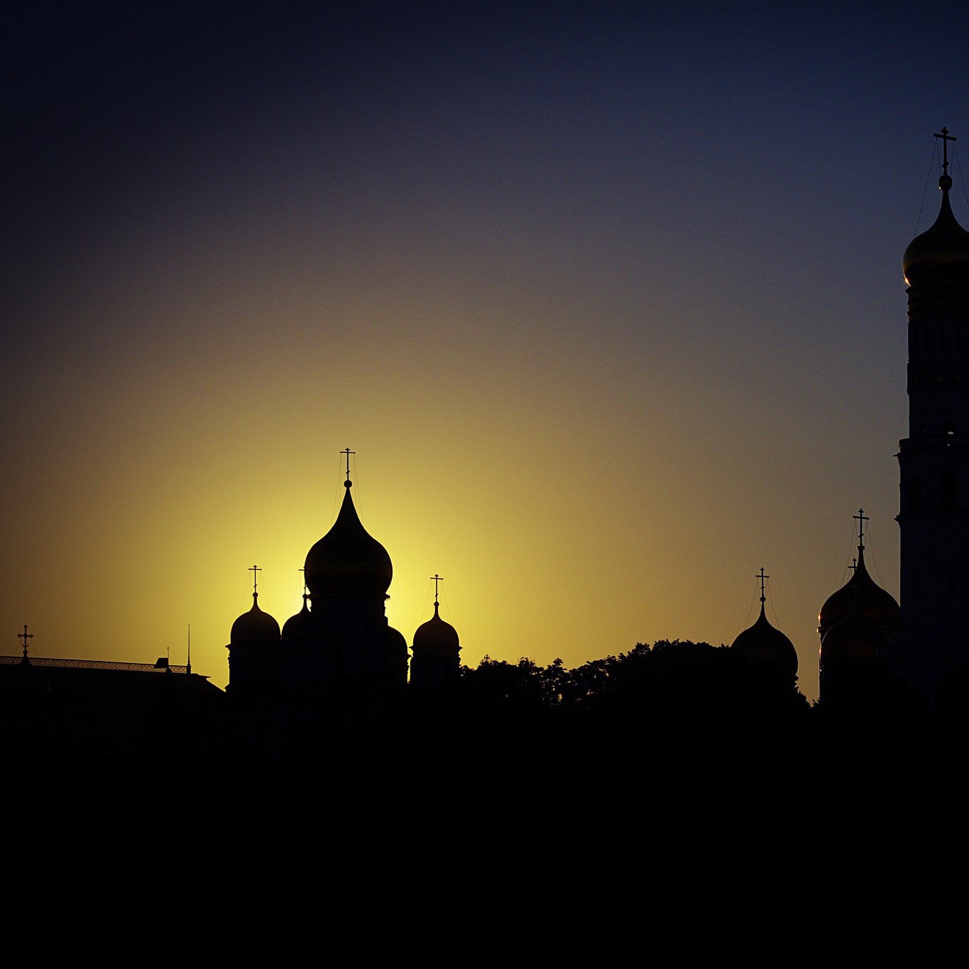 L'image présente un paysage urbain au crépuscule, où plusieurs structures architecturales se dressent en silhouette contre un ciel dégradé de couleurs. On peut apercevoir des dômes de style traditionnel, typiques de certaines églises, ainsi que des tours avec des croix au sommet. L'éclairage doux du soleil couchant crée un contraste accentué entre les ombres des bâtiments et le ciel lumineux, apportant une atmosphère calme et contemplative à la scène.