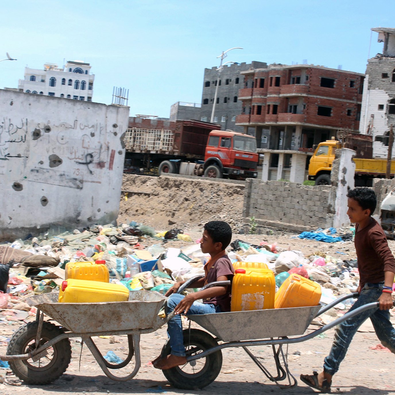 تظهر في الصورة مشهدًا مأساويًا يعكس واقعًا صعبًا. يوجد طفلان في منطقة تملأها النفايات. أحد الأطفال يدفع عربة تحتوي على دلوين، بينما يجلس الطفل الآخر في العربة. خلفهم، يمكن رؤية مباني مدمرة وأخرى قيد الإنشاء، مما يعكس تأثير النزاع أو الكوارث على هذه المنطقة. الأجواء تبدو مشمسة، لكن المشهد العام يوحي بالفقر والمعاناة.