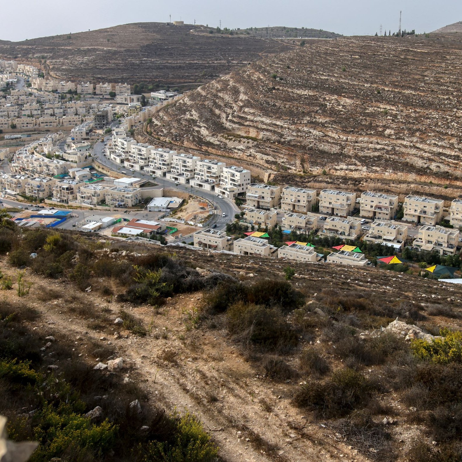 L'image montre un paysage vallonné avec des bâtiments résidentiels. On voit des maisons qui sont construites en terrasses sur les pentes de la colline, ainsi qu'une route qui serpente à travers l'urbanisation. La végétation est sparse et composée de buissons et d'herbes. Au fond, on aperçoit d'autres constructions, indiquant une zone développée et habitée. Le ciel est légèrement nuageux, ce qui donne une ambiance un peu grise à l'ensemble.
