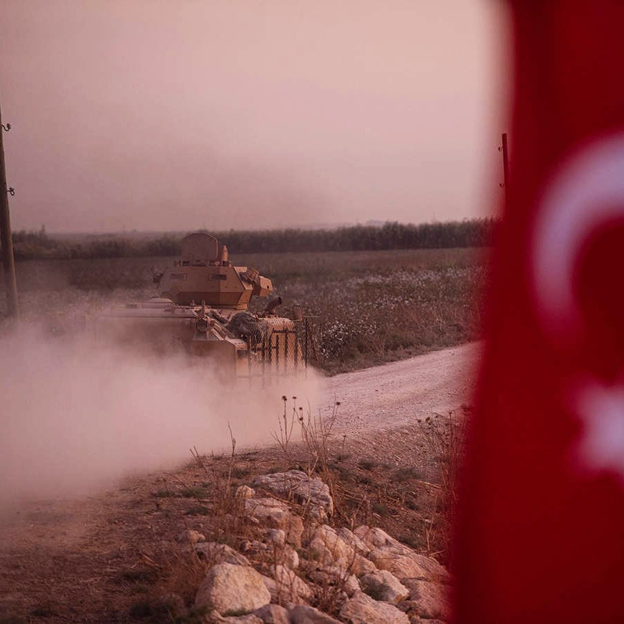 The image depicts a military tank moving along a dirt road, creating a cloud of dust. In the foreground, part of a Turkish flag is visible, featuring a white star and crescent on a red background. The landscape appears to be rural, possibly with fields or open land in the background, and the lighting suggests either dawn or dusk, giving a somewhat dramatic atmosphere to the scene.