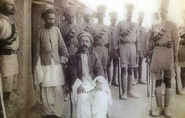 L'image montre un groupe de personnes en tenue traditionnelle, avec un homme assis au premier plan, semble-t-il en position plus détendue. Il porte des vêtements amples et un turban. Autour de lui, plusieurs hommes en uniforme militaire se tiennent debout, équipés de ceintures et de fusils, donnant une impression de formation ou de cérémonie. L'environnement semble être un lieu extérieur, probablement historique.