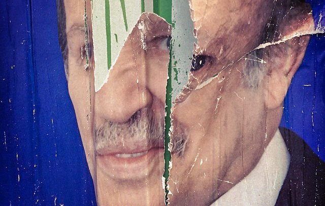 L'image montre un affichage déchiré d'un visage, probablement d'une personne publique, sur un fond bleu. Le visage est partiellement caché par des déchirures et des marques de pluie ou de peinture verte, ce qui donne un aspect usé et dégradé à l'image. Les détails du visage, comme les yeux et la bouche, sont visibles en partie, mais l'ensemble est affecté par le temps et l'usure.