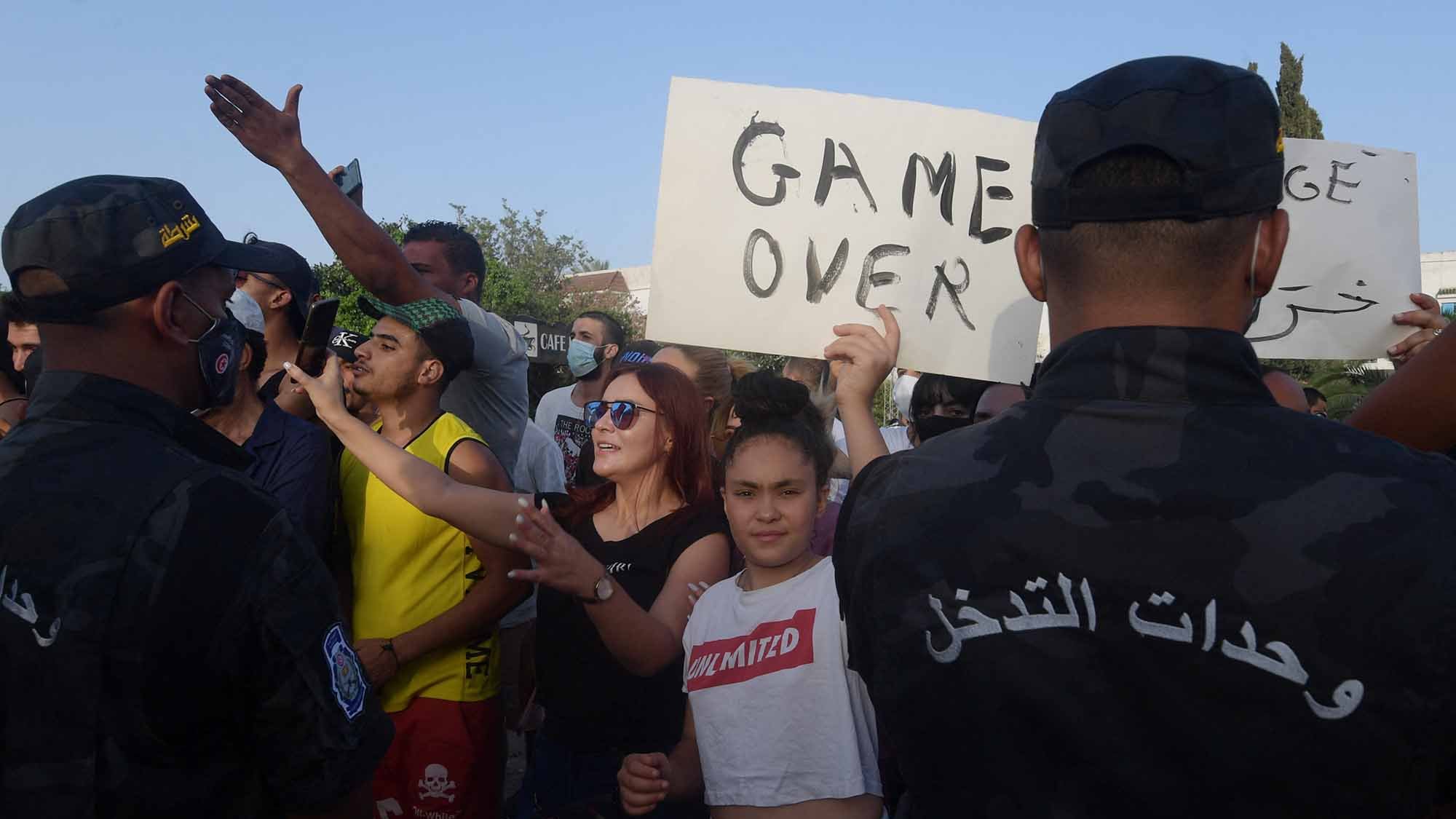 تظهر الصورة تجمعًا لعدد من الأشخاص في مُظاهرة، حيث يبدو أن هناك تبادلًا للآراء بين المحتجين والقوات الأمنية. بعض المتظاهرين يحملون لافتات، بينما يبدو آخرون يتفاعلون بشكل نشط مع الوضع. هناك تنوع في ملابس المشاركين، مما يوحي بتعدد الأعمار والجنسيات. تعكس الصورة أجواء من الحماس والاحتجاج، حيث يُظهر المحتجون رغبتهم في التعبير عن وجهات نظرهم.