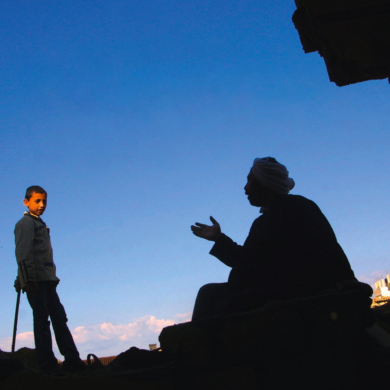 L'image montre deux silhouettes. À gauche, un garçon se tient debout, regardant vers l'autre personne. À droite, un homme est assis, en train de parler, ses bras gesticulant. Le fond est un ciel bleu, ce qui crée un fort contraste avec les silhouettes noires. L'environnement semble brut, avec des débris visibles, indiquant un cadre peut-être urbain ou une zone touchée par des événements difficiles.