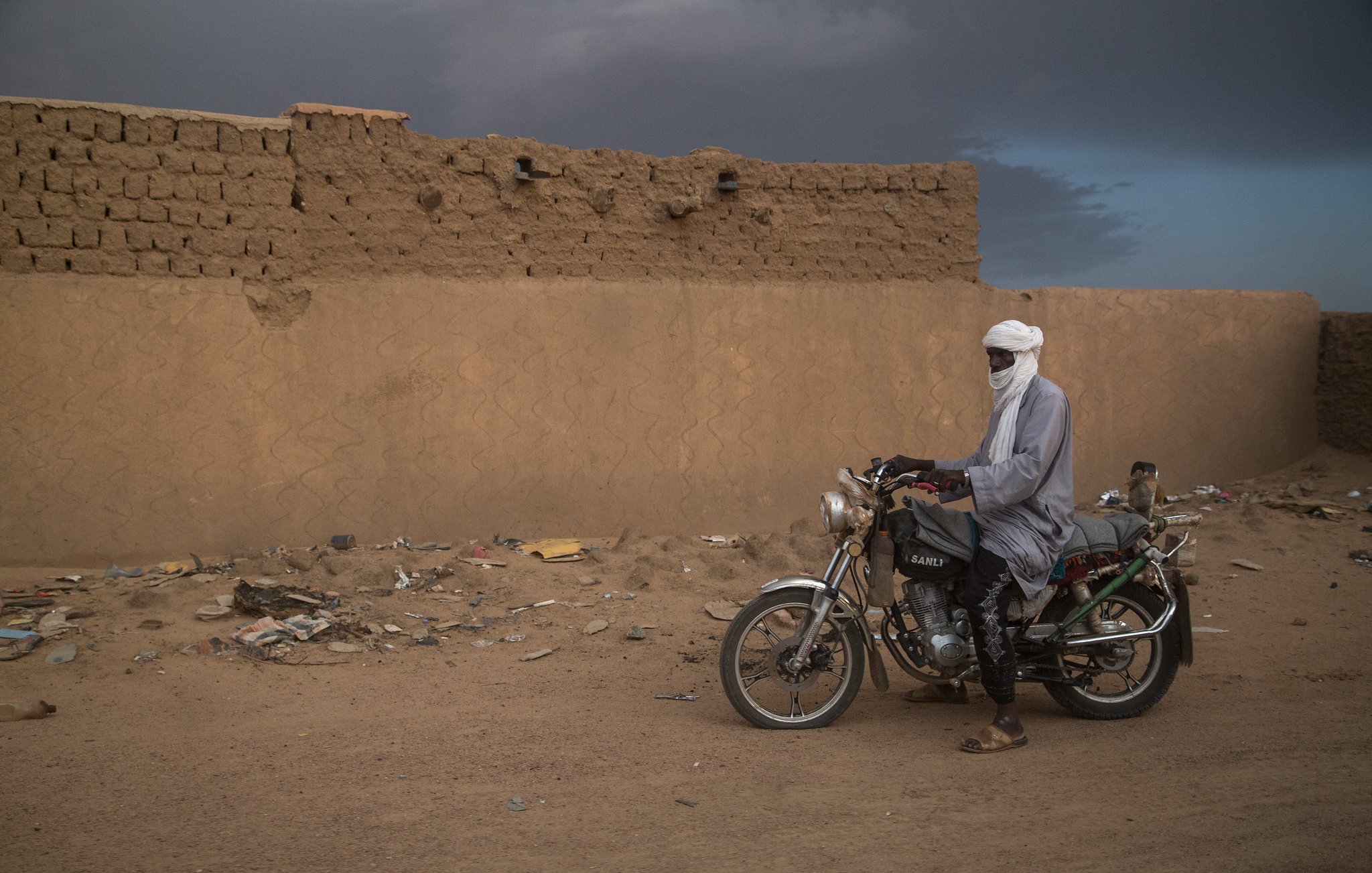 L'image montre un homme vêtu de vêtements traditionnels, avec un turban, debout à côté d'une moto. Il se trouve dans un environnement désertique, avec un mur de terre crue à l'arrière-plan. Le sol est poussiéreux et éparpillé de déchets. Le ciel semble nuageux, ajoutant une atmosphère dramatique à la scène.