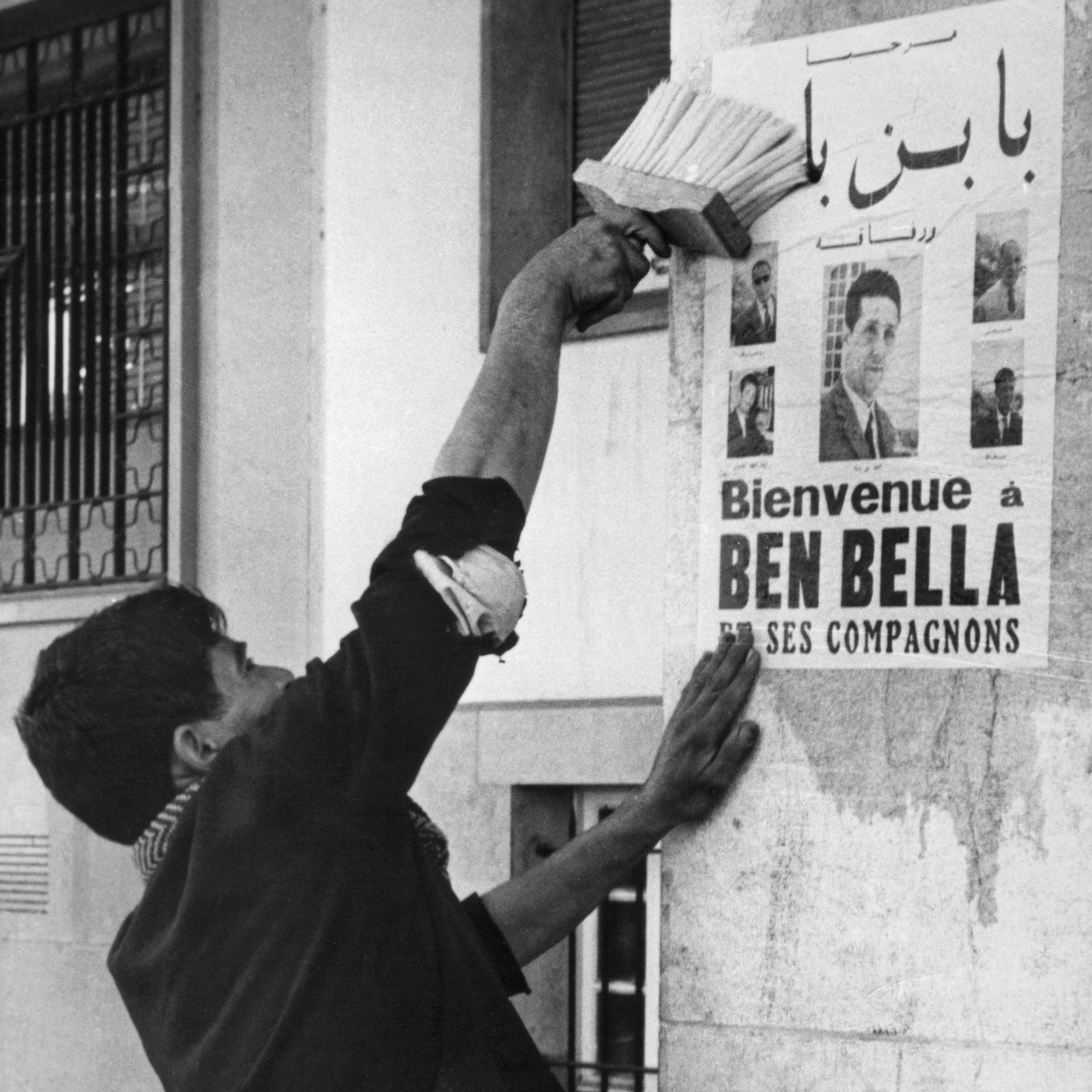 L'image montre un homme en train de coller une affiche sur un mur. L'affiche annonce un message de bienvenue à Ben Bella et ses compagnons, tout en affichant plusieurs portraits. L'homme semble concentré et utilise un outil pour bien lisser l'affiche contre la surface. L'environnement urbain suggère une période historique, probablement liée à un événement important. Les détails architecturaux et la tenue de l'homme donnent une impression de contexte social et culturel spécifique.