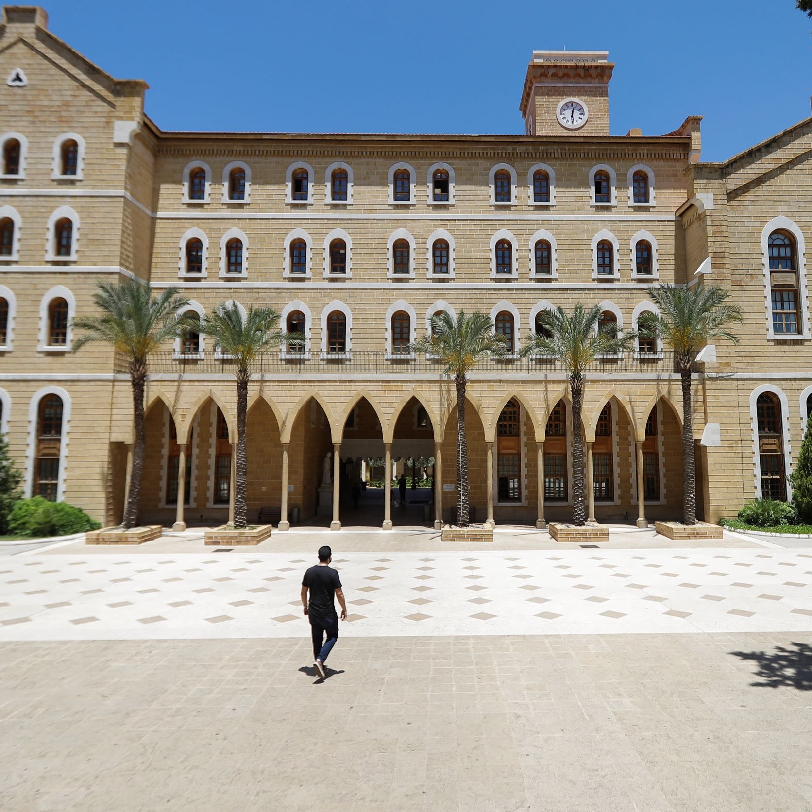 L'image montre un grand bâtiment en pierre avec une architecture traditionnelle, comportant plusieurs fenêtres à arc et des colonnes. Au premier plan, une personne marche seule sur une place pavée, tandis que des palmiers sont visibles sur les côtés. Le ciel est clair et bleu, ce qui donne une atmosphère ensoleillée. Le bâtiment semble être une institution éducative ou un lieu historique.
