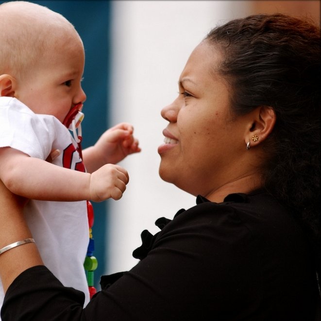 L'image montre une femme tenant un bébé dans ses bras. Le bébé, qui a une tétine, regarde la femme avec curiosité, tandis que celle-ci lui sourit. La femme porte un haut noir et a des boucles d'oreilles. L'arrière-plan est flou et coloré, suggérant un environnement en extérieur. L'atmosphère de la scène est affectueuse et chaleureuse.