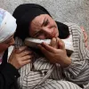 Two women in headscarves are embracing, expressing deep sorrow, with one holding a shoe.