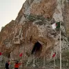 La imagen muestra un paisaje montañoso con una gran roca o acantilado en el centro, que tiene una cueva visible en su base. A su alrededor, hay varias banderas ondeando, entre las cuales algunas son de Argelia, ya que tienen un diseño distintivo. En la parte inferior de la imagen, se pueden ver algunas personas caminando y disfrutando del lugar. El cielo tiene un tono claro, posiblemente al atardecer. La escena refleja un ambiente natural y un sitio de interés cultural o histórico.