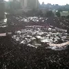 Una grande folla si raduna in una piazza, con tende e striscioni.