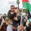 The image depicts a scene of a gathering or demonstration. A man is holding a child closely to him, showcasing a protective gesture. Around them, a crowd appears to be rallying, with some individuals raising their hands and a prominent Palestinian flag visible in the background. The atmosphere seems charged, indicating a moment of significance, possibly related to a political or social issue. The setting includes trees and a building in the background, against a cloudy sky.