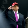 Uomo in abito scuro e cravatta viola, con berretto rosso, fa saluto militare.