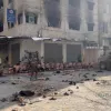 Immagine di un gruppo di persone su un marciapiede, sotto sorveglianza militare, in un contesto urbano.