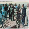 A group of military personnel and civilians gathers at a rubble site, surveying damage.