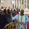 A priest in ornate vestments walks down an aisle in a church, surrounded by worshippers.