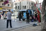 L'image montre une scène urbaine animée. Au premier plan, un homme observe une activité qui se déroule à proximité d'un van. Plusieurs personnes sont engagées dans des préparatifs, probablement pour un événement ou une manifestation. On aperçoit des drapeaux, probablement le drapeau libanais, qui ajoutent une touche de couleur à la scène. En arrière-plan, on distingue des bâtiments typiques, évoquant une ambiance de ville. L'atmosphère semble à la fois dynamique et communautaire.