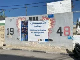 L'image montre un mur mural avec une grande affiche au centre. L'affiche est en arabe et en anglais, déclarant que les services de l'UNRWA (l'Agence des Nations Unies pour les réfugiés de Palestine) sont un droit jusqu'au retour. Le mur est orné de graffiti, incluant des chiffres "19" et "48", probablement en référence à des événements historiques liés aux réfugiés palestiniens. L'environnement semble être un espace urbain, peut-être dans un camp de réfugiés, car le mur et le décor suggèrent un contexte communautaire.