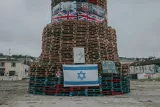 L'image montre une structure imposante construite avec des palettes en bois, ornée de différents drapeaux, y compris un drapeau israélien. Il y a également des affiches et des inscriptions sur des panneaux, et l'environnement semble être une zone urbaine avec des bâtiments en arrière-plan. Le ciel est nuageux, ce qui donne une atmosphère sombre à la scène.