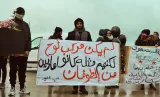 L'image montre un groupe de manifestants, certains portant des masques et des vêtements sombres, rassemblés sous un ciel nuageux. Au centre, un homme tient une grande pancarte ornée de slogans en arabe. La pancarte semble exprimer un message fort, et plusieurs personnes autour de lui sont également munies de bouquets de fleurs. L'atmosphère générale de l'image reflète un moment de protestation ou de commémoration.