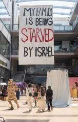 Une grande banderole dénonce la souffrance d'une famille en lien avec Israël. Des personnes marchent.