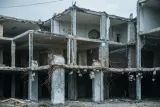 L'image montre un bâtiment en ruines, avec une structure incomplète. Les murs sont partiellement effondrés et on peut voir des barres d'armature exposées ainsi que des débris éparpillés au sol. L'ensemble a l'air abandonné et en décomposition, avec un ciel nuageux en arrière-plan, ce qui renforce l'atmosphère de désolation.