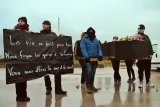 L'image montre un groupe de manifestants tenant des pancartes et un cercueil. Les pancartes affichent des messages politiques sur le droit à la vie et la fuite des guerres. L'ambiance semble sombre et sérieuse, avec des personnes portant des vêtements chauds, ce qui suggère des conditions météorologiques difficiles. L'un des manifestants porte un bouquet de fleurs, ajoutant une dimension symbolique à la protestation.