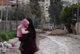 L'image montre une femme portant un enfant, marchant sur une rue poussiéreuse et inégale, avec des obstacles en terre et des déchets sur le sol. En arrière-plan, on aperçoit des véhicules militaires et des bâtiments. L'ambiance est sombre et témoigne d'un environnement urbain difficile.