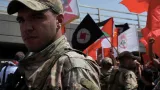 Un soldat en uniforme, avec des manifestants en arrière-plan portant des drapeaux colorés.