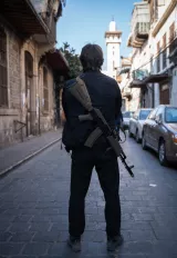 Un homme de dos se tient sur une rue pavée, il porte un gilet noir et porte une arme sur son épaule. En arrière-plan, on peut voir des bâtiments anciens et un minaret. L'atmosphère est calme, mais l'image dégage une certaine tension.