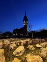 L'image montre une scène nocturne avec un ciel bleu foncé. En arrière-plan, on aperçoit une grande tour avec une horloge, probablement un bâtiment historique. Au premier plan, il y a un décor de rochers et d'herbes, ainsi qu'une clôture en métal. L'ensemble donne une atmosphère calme et mystérieuse, accentuée par l'éclairage subtil du bâtiment et l'obscurité environnante.