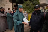 L'image montre une file de personnes, principalement des femmes en hijab, attendant. Au premier plan, une femme en tenue verte tient un paquet dans ses mains, en interaction avec un homme vêtu d'un manteau noir. L'environnement est urbain et chargé de végétation.