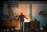 L'image montre un homme debout devant un feu, avec un décor mural en arrière-plan représentant un paysan au travail. Le panneau au-dessus indique qu'il s'agit des bureaux de l'UNRWA, un programme de secours et de travaux pour les réfugiés palestiniens. La scène évoque un contexte de vie difficile, probablement lié à des problématiques sociales ou politiques. L'éclairage du feu crée une atmosphère intense et dramatique.