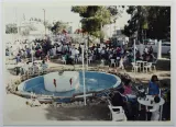 L'image montre un parc animé avec de nombreuses personnes. Au centre, il y a une fontaine entourée d'eau et de quelques fauteuils. Des groupes de personnes sont assis à des tables, discutant et profitant de l'ambiance. En arrière-plan, on aperçoit des aires de jeux pour enfants. L'espace est ombragé par des arbres, ce qui ajoute une atmosphère conviviale. Il semble s'agir d'un lieu de rassemblement populaire, rempli d'activités et de vie.