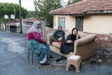 L'image montre trois femmes assises sur un canapé dans un environnement rural. Leurs tenues sont traditionnelles et elles semblent discuter ensemble. Une femme est assise sur une chaise, tandis que les deux autres sont sur le canapé. En arrière-plan, on peut voir des maisons avec des toits en tuiles, ainsi que des arbres et des antennes parabolique. L'ambiance semble tranquille et conviviale.