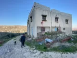 L'image montre un bâtiment abandonné, avec une structure en béton et des fenêtres grillagées. Il y a des traces de peinture rouge sur le mur extérieur. À côté du bâtiment, une personne marche avec un chien. Le sol est couvert de gravier et de végétation, et à l'arrière-plan, on aperçoit des collines et un village. L'éclairage semble naturel, suggérant que la photo a été prise en plein jour.