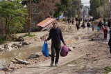 L'image montre un homme marchant sur un chemin inégal, portant deux sacs en plastique. En arrière-plan, on aperçoit d'autres personnes se déplaçant, ainsi que des débris et des constructions inachevées. Le paysage semble désordonné et en chantier.